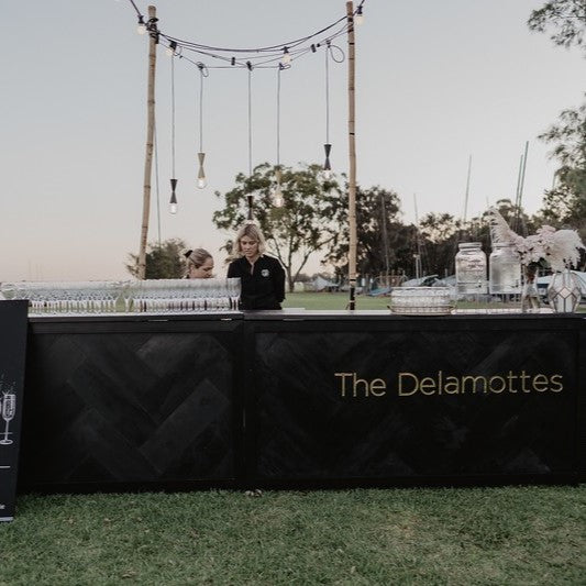 Two people stand behind a black bar counter labeled "The Delamottes" outdoors. The bar is set up on a grassy area with a few decorative elements and glasses lined up. Trees and a clear sky are in the background.