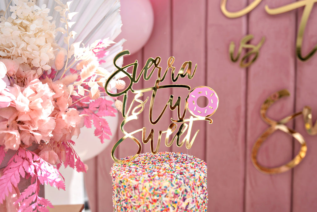 A colorful birthday cake topped with rainbow sprinkles features a gold "Sierra is Two Sweet" cake topper. The background is decorated with pink floral arrangements and a pink wooden wall.