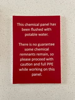 **A red, custom-designed Etched Design Acrylic Business Sign created with Traffolyte features white text that reads: "This chemical panel has been flushed with potable water. There is no guarantee some chemical remnants remain, so please proceed with caution and full PPE while working on this panel." Precision laser engraving ensures the text's clarity. The sign is displayed on a light-colored surface.**