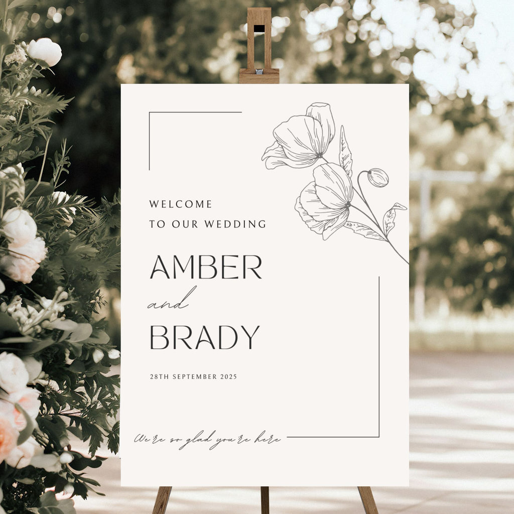 An acrylic wedding sign from Etched Design titled "Wedding Welcome Sign | Amber & Brady" is showcased on an easel, displaying the message "Welcome to our wedding Amber and Brady, 28th September 2025. We're so glad you're here." The sign features minimalist floral illustrations and is surrounded by greenery and flowers in an outdoor setting.