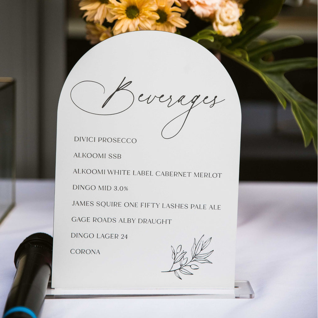 A "Wedding Beverage Menu | Botanical" by Etched Design is elegantly displayed on an acrylic table sign with a cursive font at the top. Decorative flowers in the background add a charming touch. The menu lists various alcoholic beverages including prosecco, wines, beer, and Corona. A microphone is placed next to this custom-designed wedding decor piece.