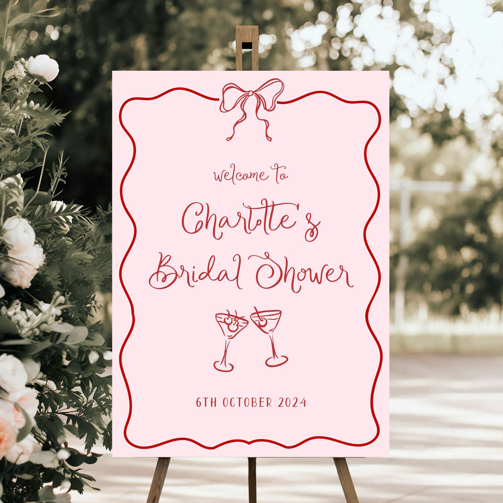 An elegant personalised bridal shower sign by Etched Design stands on an easel, reading "Welcome to Charlotte's Bridal Shower, 6th October 2024" in sophisticated red lettering. This acrylic bridal shower welcome sign features a pink background with a decorative red border and a ribbon illustration at the top, complemented by two clinking cocktail glasses at the bottom.