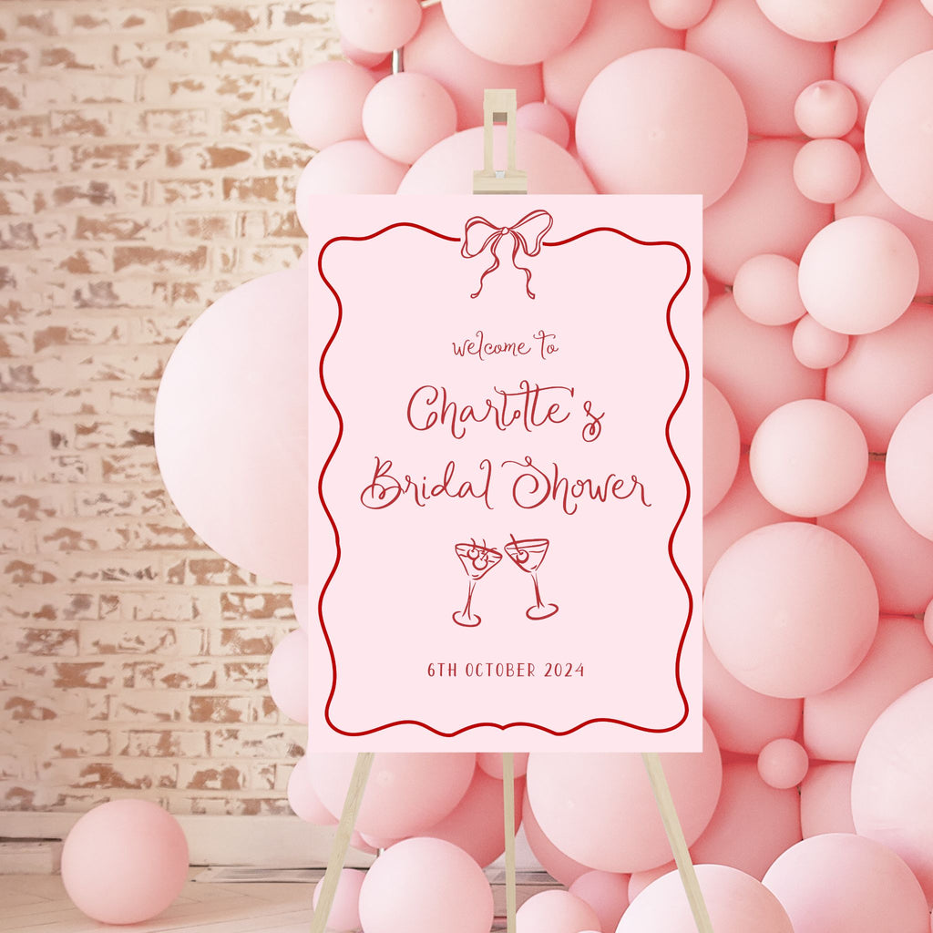 A personalized bridal shower sign from Etched Design sits on an easel amidst a backdrop of pink balloons, featuring the product name "Bridal Shower Welcome Sign | Charlotte." The sign reads, "Welcome to Charlotte's Bridal Shower, 6th October 2024" in elegant pink script. Above the text is a charming illustration of a pink bow, and below it are two clinking cocktail glasses.