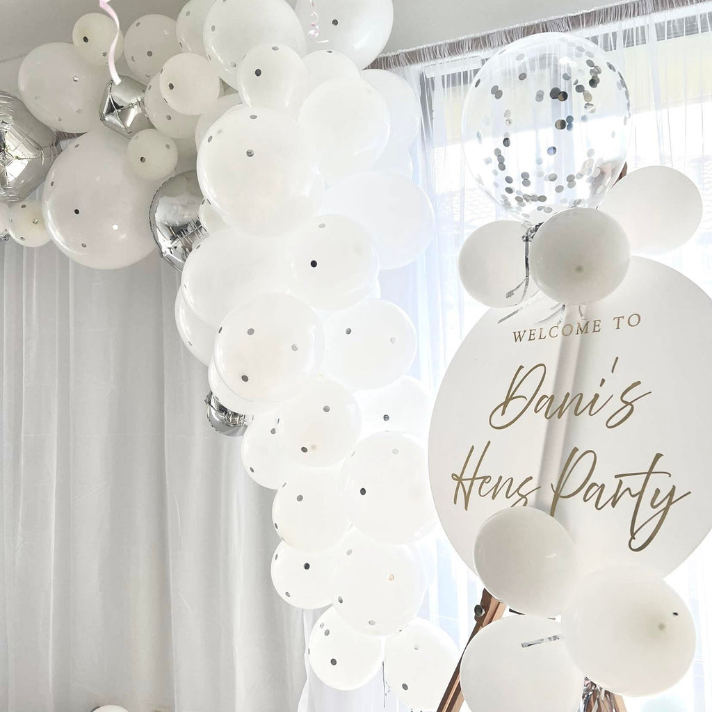 A decorative setup showcases a white balloon arch featuring a blend of solid white, clear polka-dotted, and metallic silver balloons. A Custom Round Welcome Sign by Etched Design elegantly reads "Welcome to Dani's Hens Party" in beautiful gold script. The backdrop is composed of light, sheer curtains.