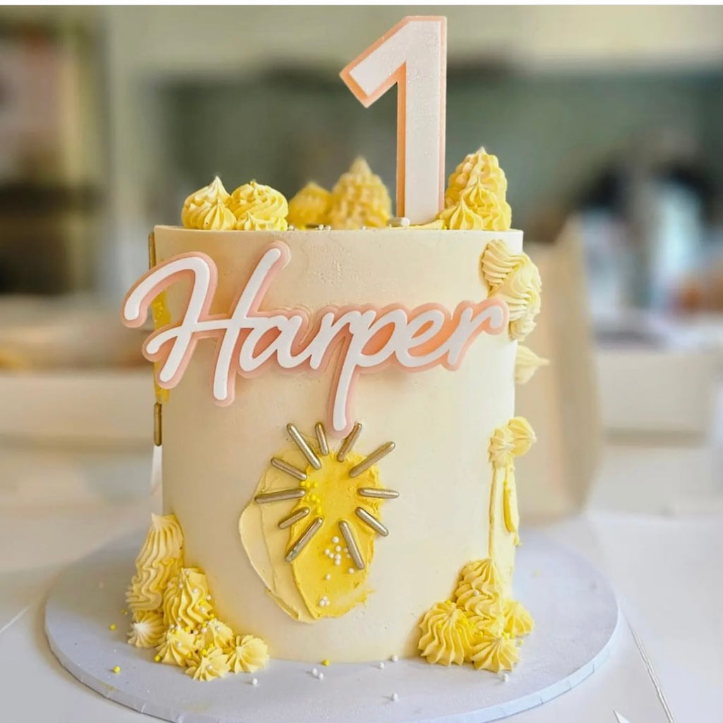 A tall white birthday cake, decorated with yellow frosting and adorned with piped rosettes and sun-themed accents, features a charming "1" cake topper from the Etched Design's Name & Age Cake Topper Set. The name "Harper" is written in pink on top of the cake, which sits elegantly on a white round board.