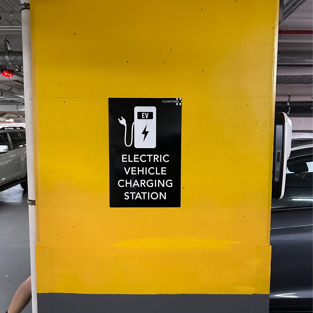 A yellow and grey wall in a parking garage features an Acrylic Business Sign | Traffolyte from Etched Design, custom-designed with a laser-etched black surface displaying a white electric vehicle plug and battery icon. The sign reads "ELECTRIC VEHICLE CHARGING STATION," indicating the presence of a charging facility for electric vehicles nearby.