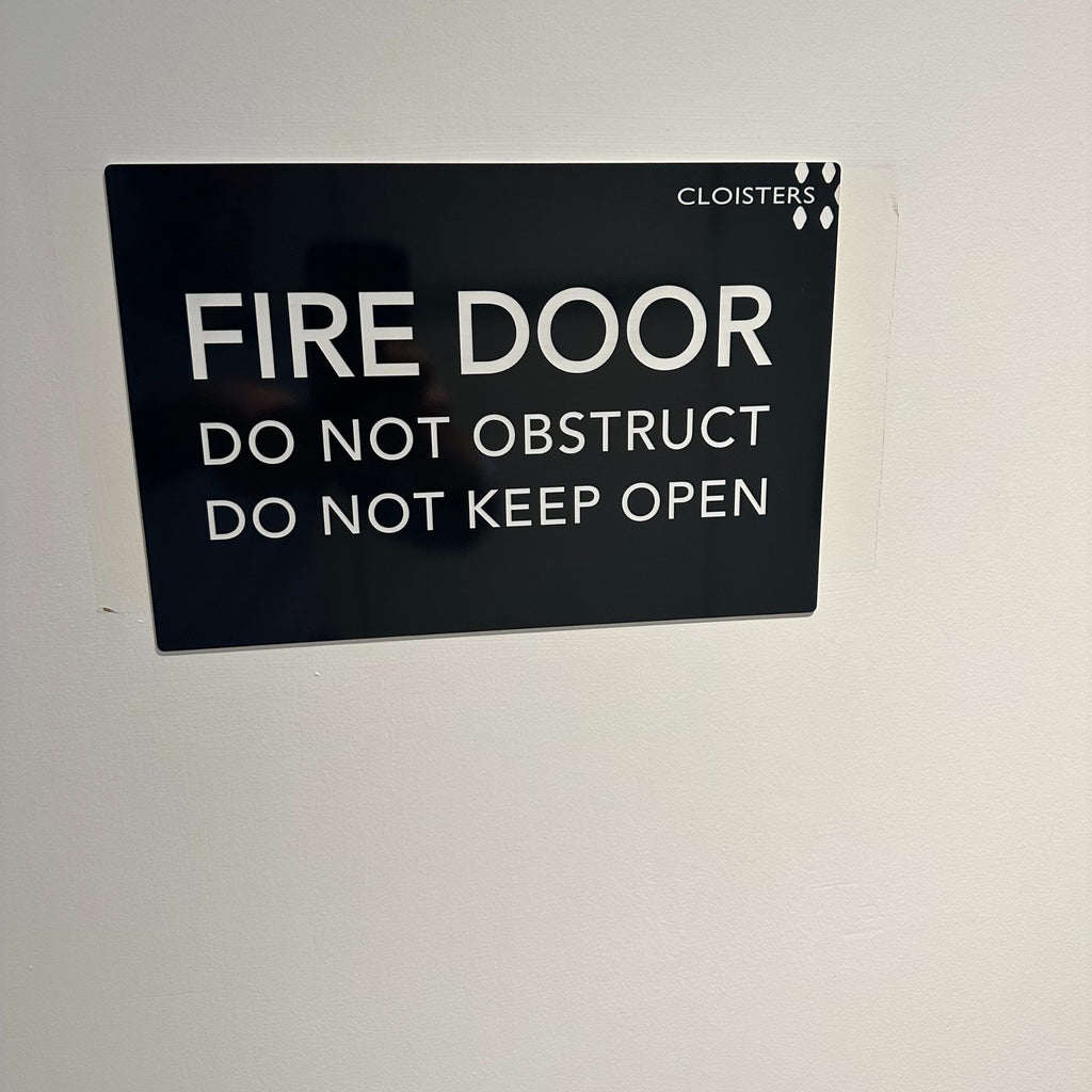 A black Acrylic Business Sign (Traffolyte) from Etched Design with white text on a wall reads: "FIRE DOOR. DO NOT OBSTRUCT. DO NOT KEEP OPEN." The laser-etched sign also includes the word "CLOISTERS" in the upper right corner.