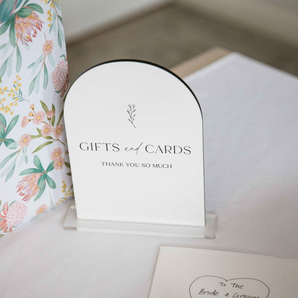 The "Gifts and Cards" Botanical sign by Etched Design is an elegant, white acrylic table sign with an arched top and a delicate floral illustration. Displayed on a clear stand, it offers gratitude to guests. It is set on a table adorned with wedding decorations, featuring a floral gift bag and a partially visible card in the corner.