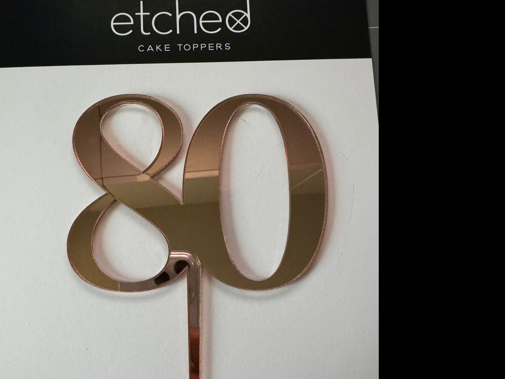 A rose gold mirrored acrylic cake topper in the shape of the number "80" against a white background. The packaging label at the top reads "Etched Design CAKE TOPPERS." This elegant piece, known as the 80th Cake Topper, is perfect for clearance items and adds a touch of sophistication to any celebration.