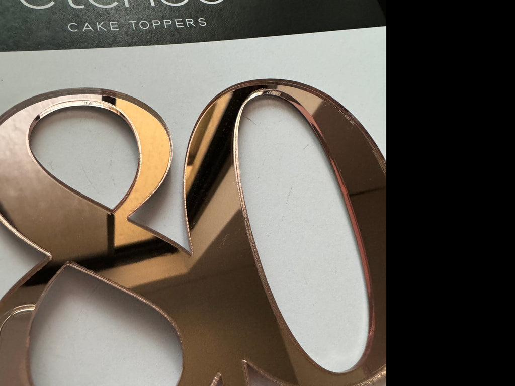 Close-up image of an Etched Design 80th Cake Topper, made from rose gold mirrored acrylic and shaped like the number "80." The background includes part of a dark label with the text "cake toppers.