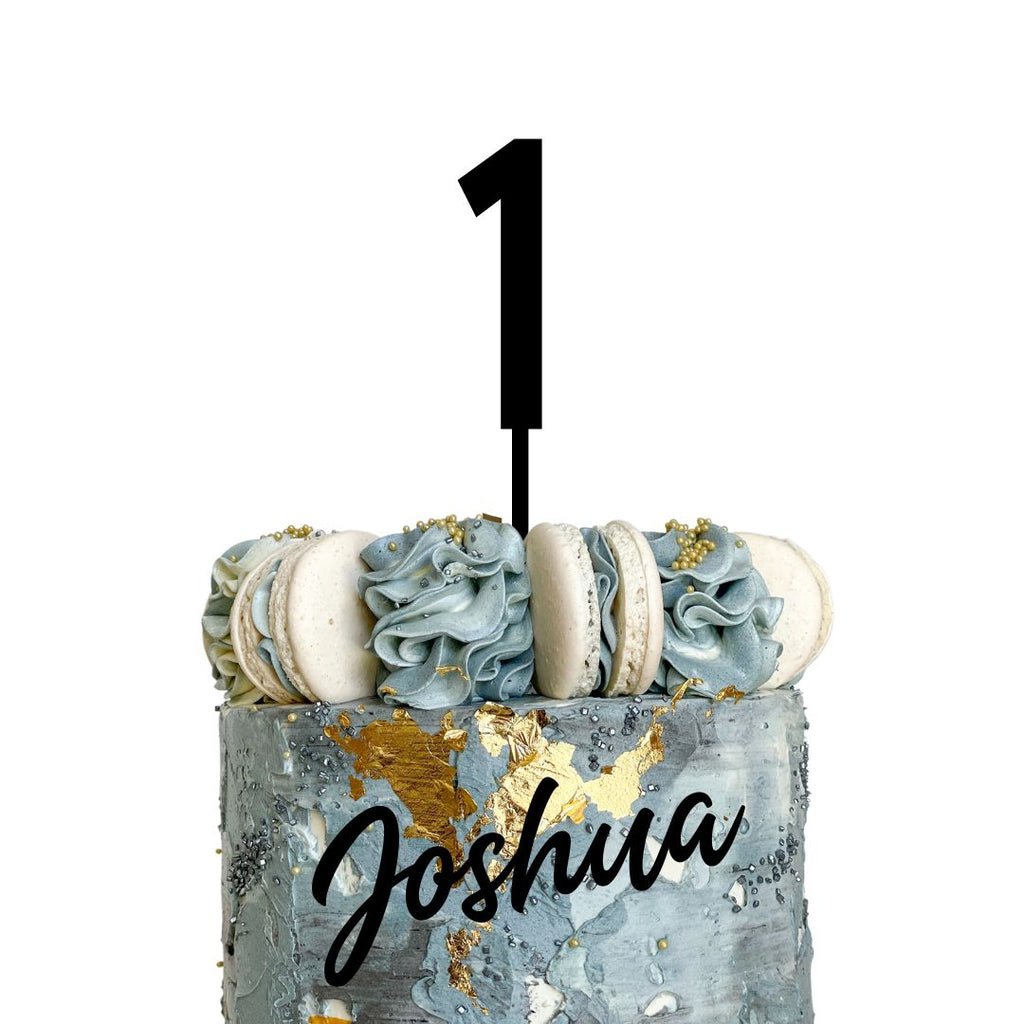 A decorated double layered cake with a gold and marble texture, featuring Etched Design's Name & Age Cake Topper Set showcasing a large number "1" topper. The cake is adorned with blue-grey rosettes and macarons, and includes the name "Joshua" written in black cursive on the side for personalized decoration.