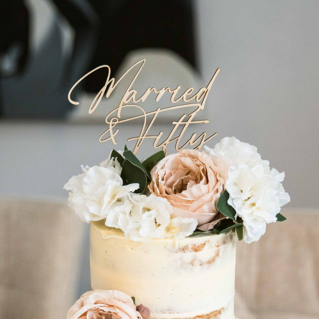 A semi-naked wedding cake adorned with white and blush pink flowers, featuring an Etched Design custom cake topper that reads "Married & Flirty." The background is softly blurred, highlighting the bespoke design of the cake as the central focal point.