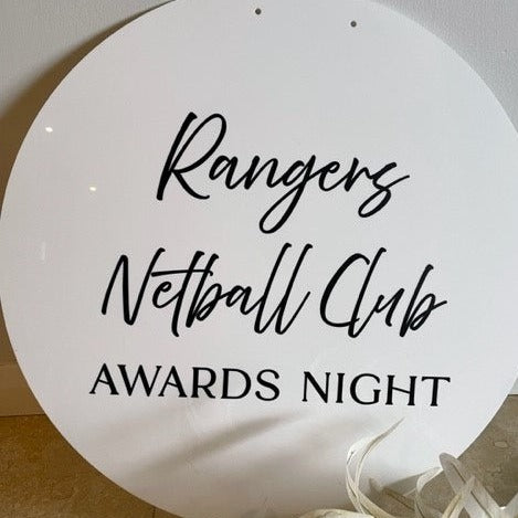 A Custom Round Welcome Sign by Etched Design, featuring the custom text "Rangers Netball Club Awards Night" in black cursive and bold fonts on a white acrylic circular design. This personalized event decor is set against a light background, adorned with white ribbons at the bottom edge.