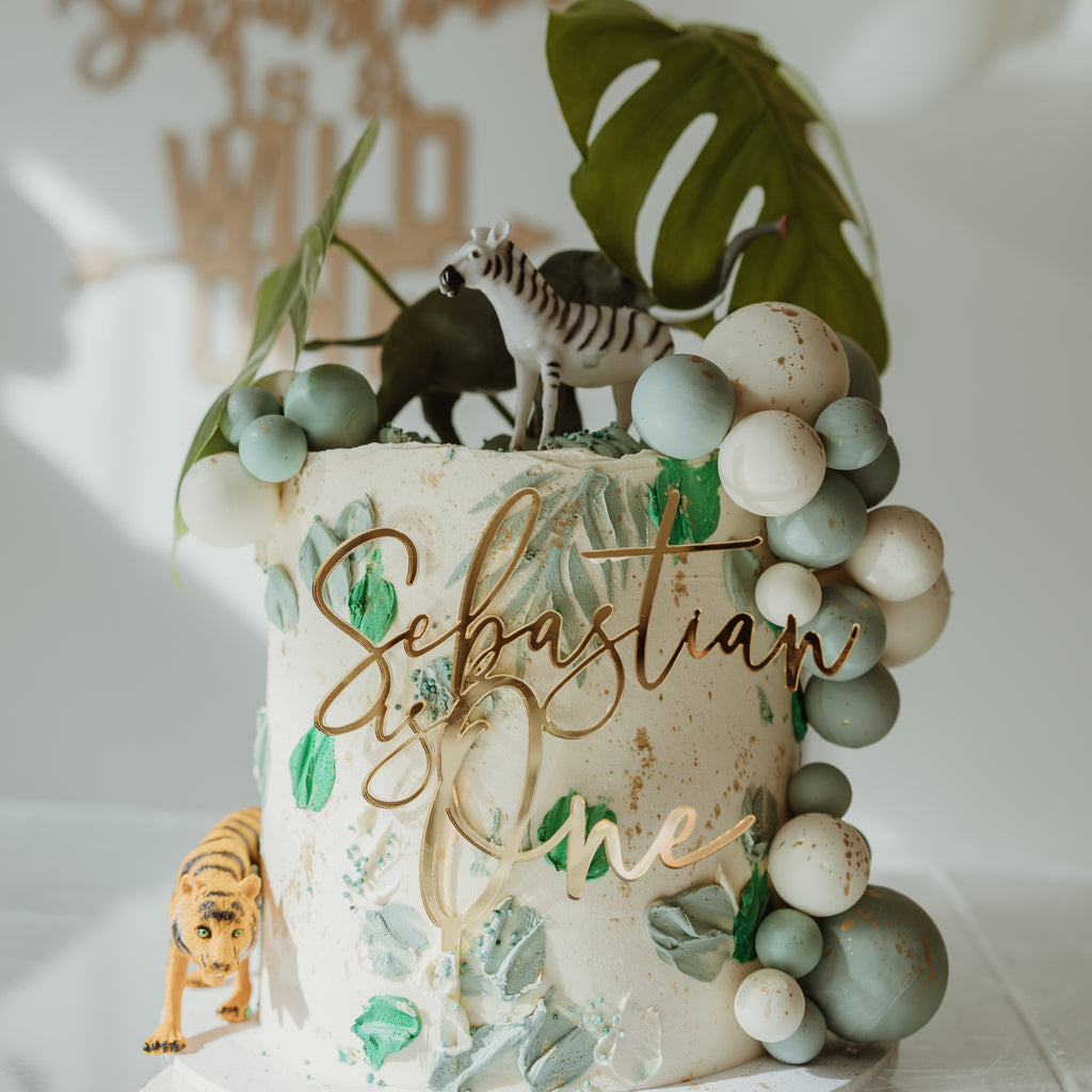 A Cake Fropper | Single Layer by Etched Design, jungle-themed and decorated with green leaf, zebra, tiger, and dinosaur figurines, along with green and white balloon-like icing. An acrylic cake plaque reads "Sebastian is One." The personalized cake decoration is placed on a white surface with a blurred background.