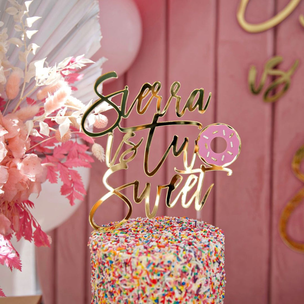 A vibrant cake adorned with rainbow sprinkles is topped with Etched Design's Custom Cake Topper reading "Sierra is Two Sweet," with a pink donut illustration replacing the 'o' in "Two". The background features pastel decorations and a wooden wall with additional text.