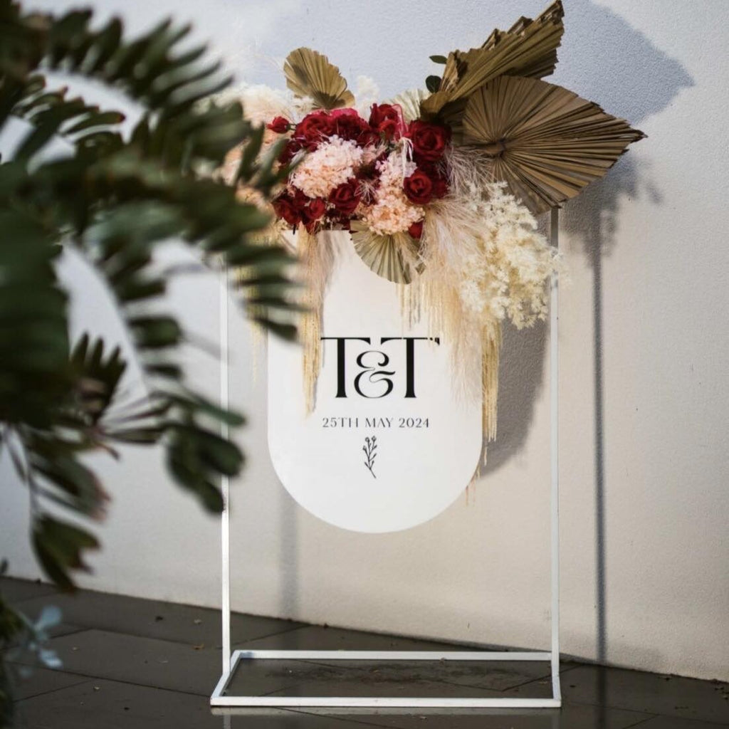 A personalized Engagement Welcome Sign by Etched Design featuring the initials "T&T" and the date "25th May 2024." This minimalist decor for weddings showcases a white background elegantly adorned with red and pink flowers, pampas grass, and large dried palm leaves. The welcoming foreground is framed by leafy plants.