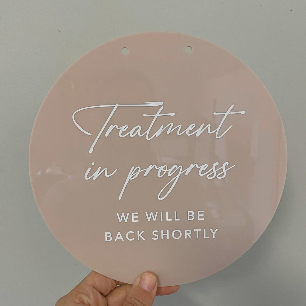 A hand holds the Etched Design Custom Open/Closed Door Sign - Botanicals against a plain, light gray background. The round, blush-colored acrylic sign features white script text that reads "Treatment in progress" and uppercase letters below stating "We will be back shortly.