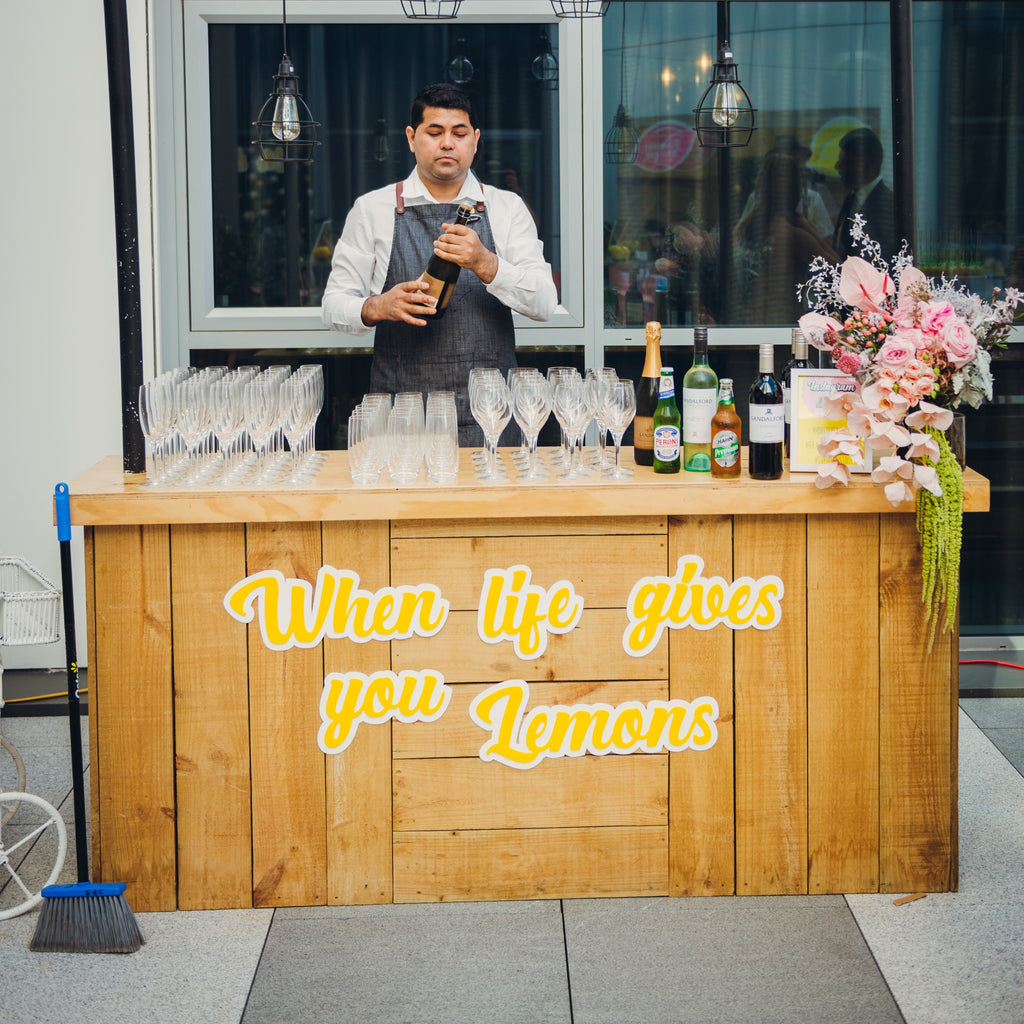 A bartender stands behind a wooden bar, which showcases an Etched Design 3D Bar Sign that reads "When life gives you Lemons" in vibrant yellow script. The bar is equipped with multiple wine glasses, bottles, and a beautiful floral arrangement. In the background, there are windows and pendant lights, while a broom leans nearby.