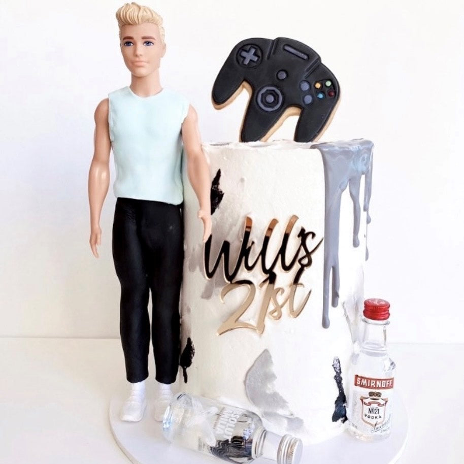 A decorative Cake Fropper | Single Layer by Etched Design, featuring the text "Will's 21st" on an acrylic cake plaque. The cake is adorned with a black game controller on top, and beside it stands a male doll dressed in a light blue shirt and black pants. Two small bottles of vodka are placed at the base of the cake.