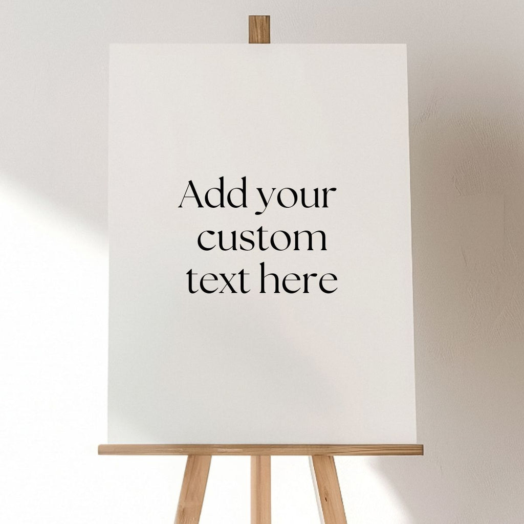 A Custom Acrylic Welcome Sign by Etched Design is positioned on a wooden easel against a plain wall. The elegant black font on the blank white canvas invites, "Add your custom text here," making it perfect for any personalized event.