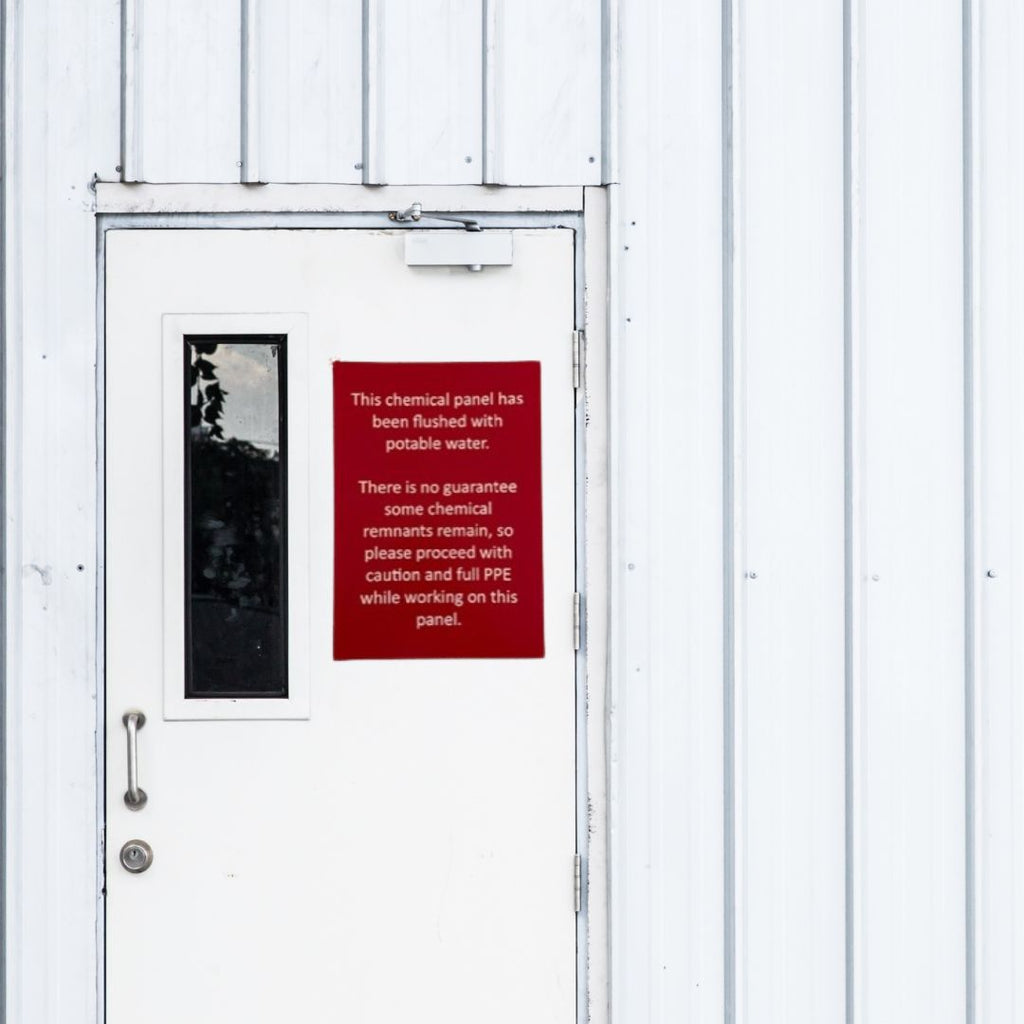 A white industrial door with a vertical narrow window features a red, custom-designed acrylic sign by Etched Design. The sign, crafted using precision laser engraving, reads: "This chemical panel has been flushed with potable water. There is no guarantee some chemical remnants remain, so please proceed with caution and full PPE while working on this panel.