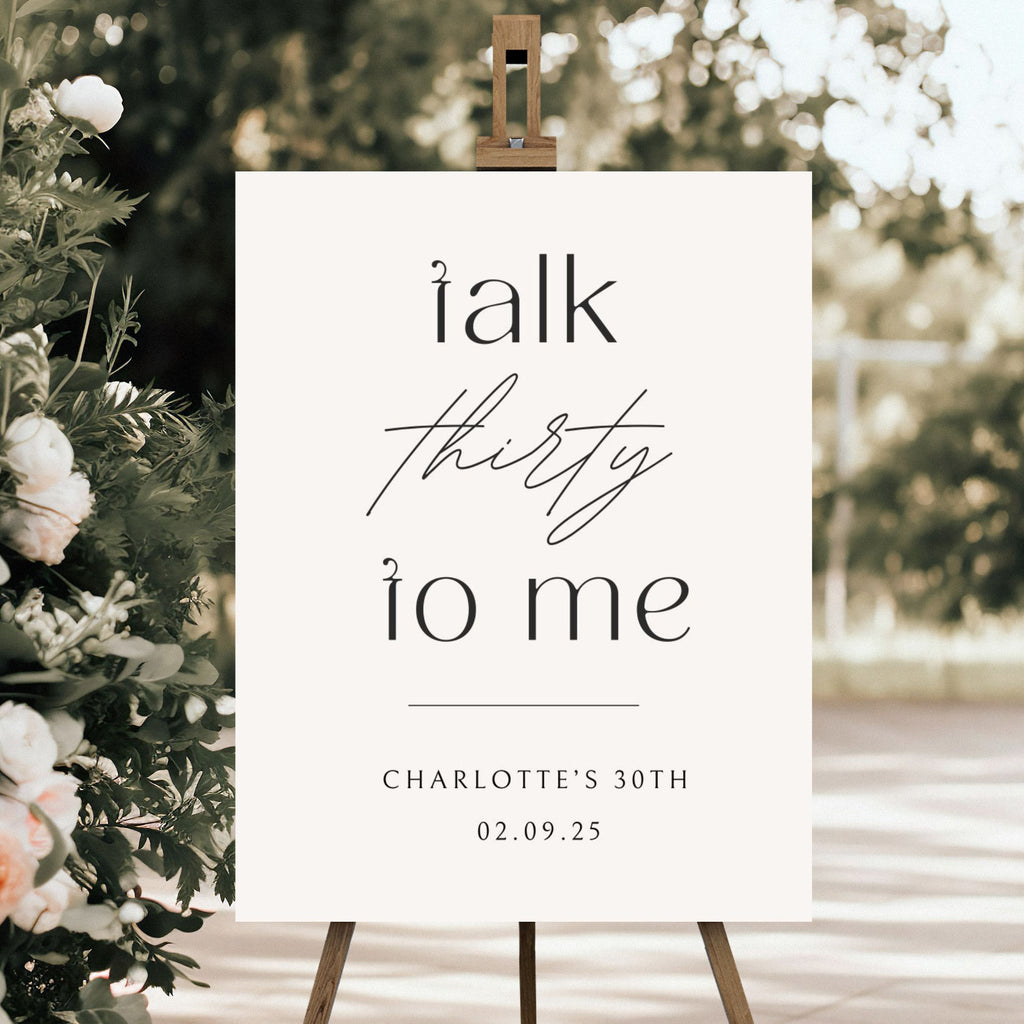 A personalised welcome sign on an easel from Etched Design, named "Welcome Sign | Talk Thirty To Me," displays the text "Talk thirty to me" in elegant fonts. Below, it reads "CHARLOTTE'S 30TH" and "02.09.25". The background features a blurry outdoor setting with greenery, with floral arrangements visible on the left side of the image.