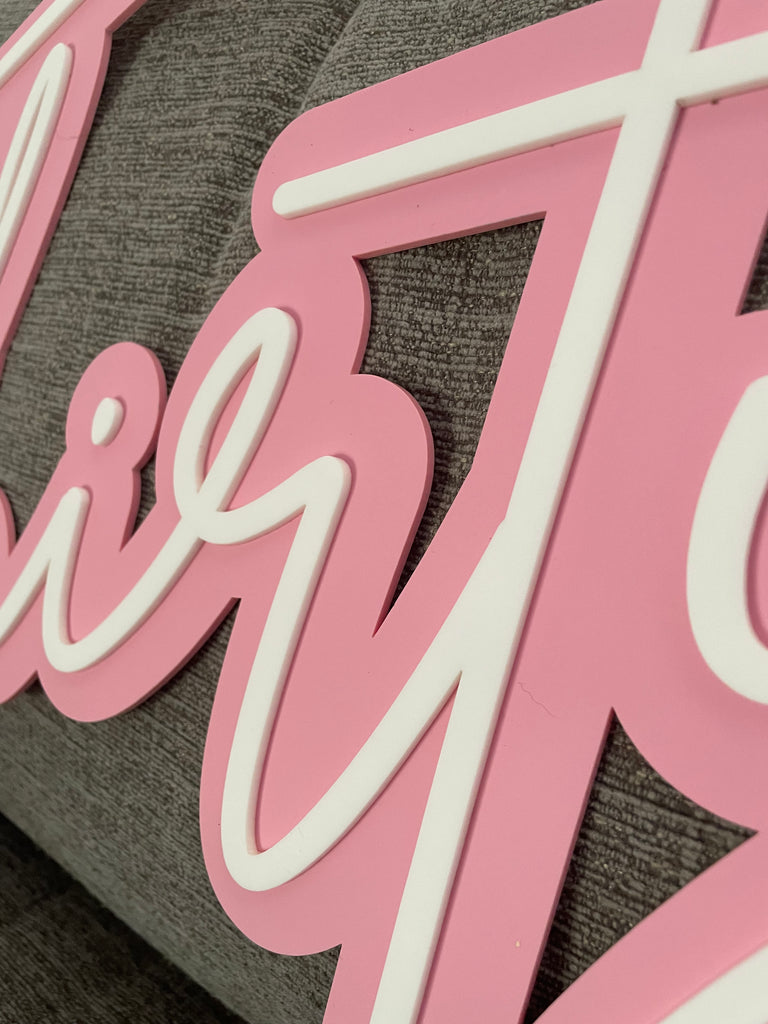 Close-up of a pink and white Name Plaque | 3D by Etched Design against a gray fabric background. The design features modern script font in cursive lettering, though the words are not fully visible in the image.