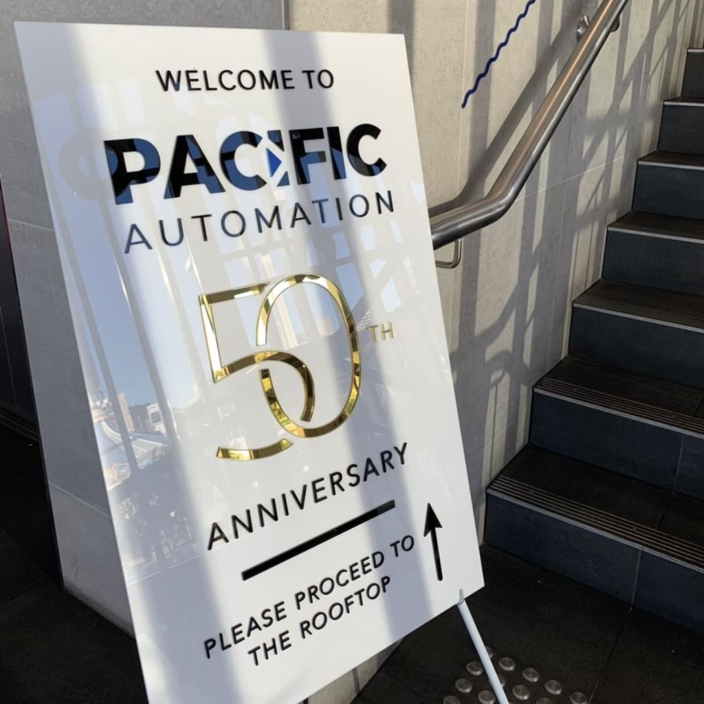An Etched Design Corporate Welcome Sign - Acrylic, custom-designed with "Welcome to Pacific Automation 50th Anniversary" and a directional arrow pointing to the rooftop, stands near a stairway. The black and gold laser-cut sign, lit by sunlight, casts reflections on its shiny surface.