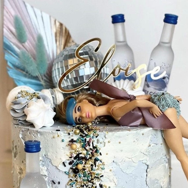 The decorated cake is highlighted by a bespoke "Name Cake Topper" from Etched Design, featuring a doll lying on its side with a brown ribbon and blue headband. The personalized topper is adorned with colorful beads, decorations such as a disco ball, feathers, and two bottles. The name "Eloise" is prominently displayed at the top, making it a truly personalized masterpiece.