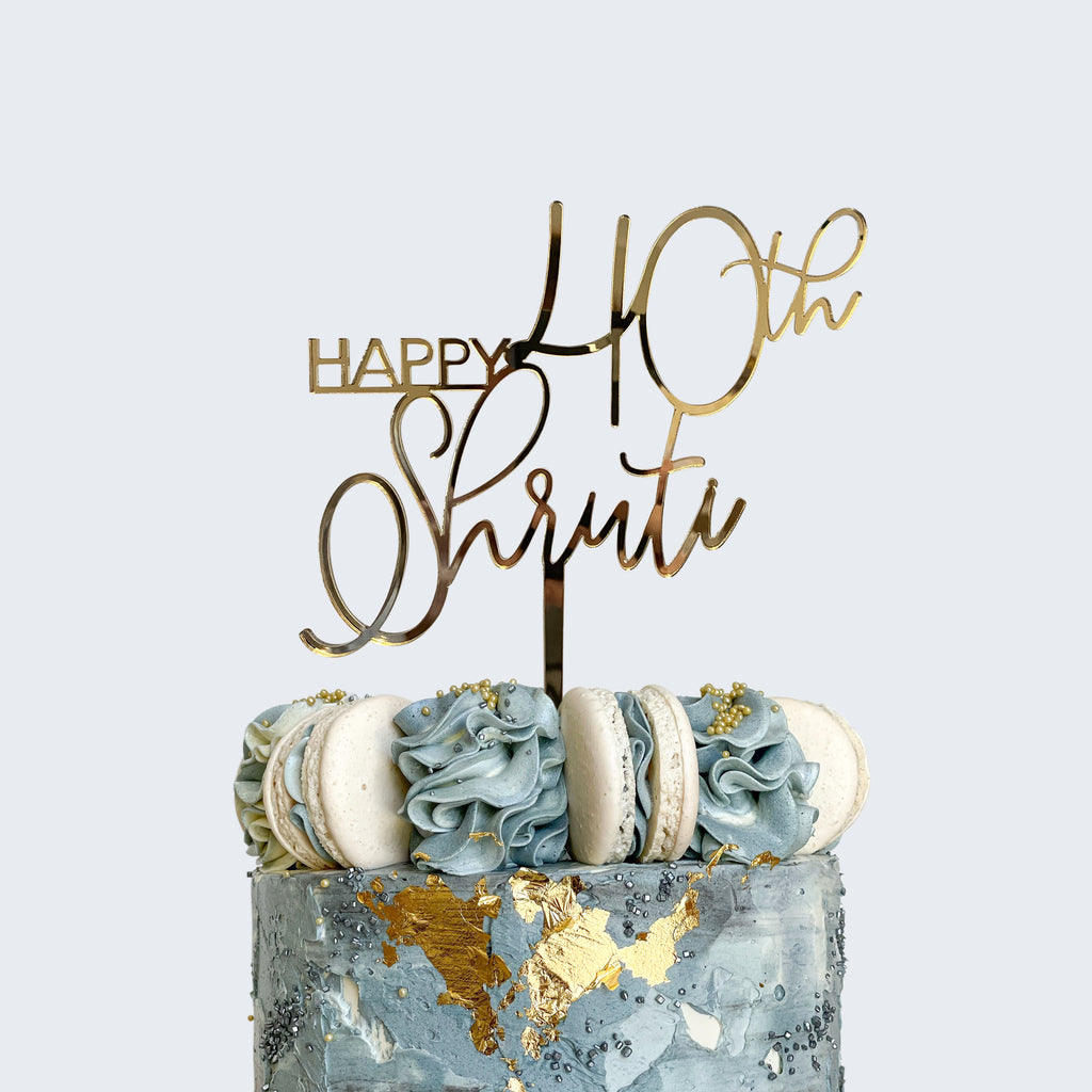 A decorative, personalized "Happy 40th Shruti" Script Birthday Cake Topper from Etched Design graces the birthday cake, which boasts blue and white frosting swirls, gold accents, and macarons. The backdrop is a plain, light gray color.