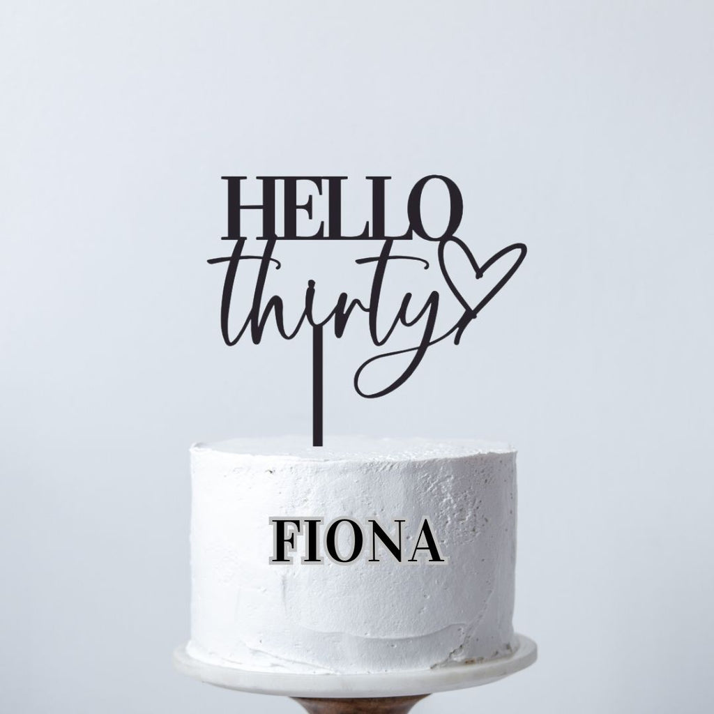 A white cake with a smooth, frosted finish stands on a wooden cake stand. The custom cake decoration reads "Hello forty" in cursive, featuring a heart symbol. Bold, black letters spell out the name "Fiona" on the front of the milestone birthday cake. The background is plain white.

Product shown: Hello Forty Cake Topper by Etched Design