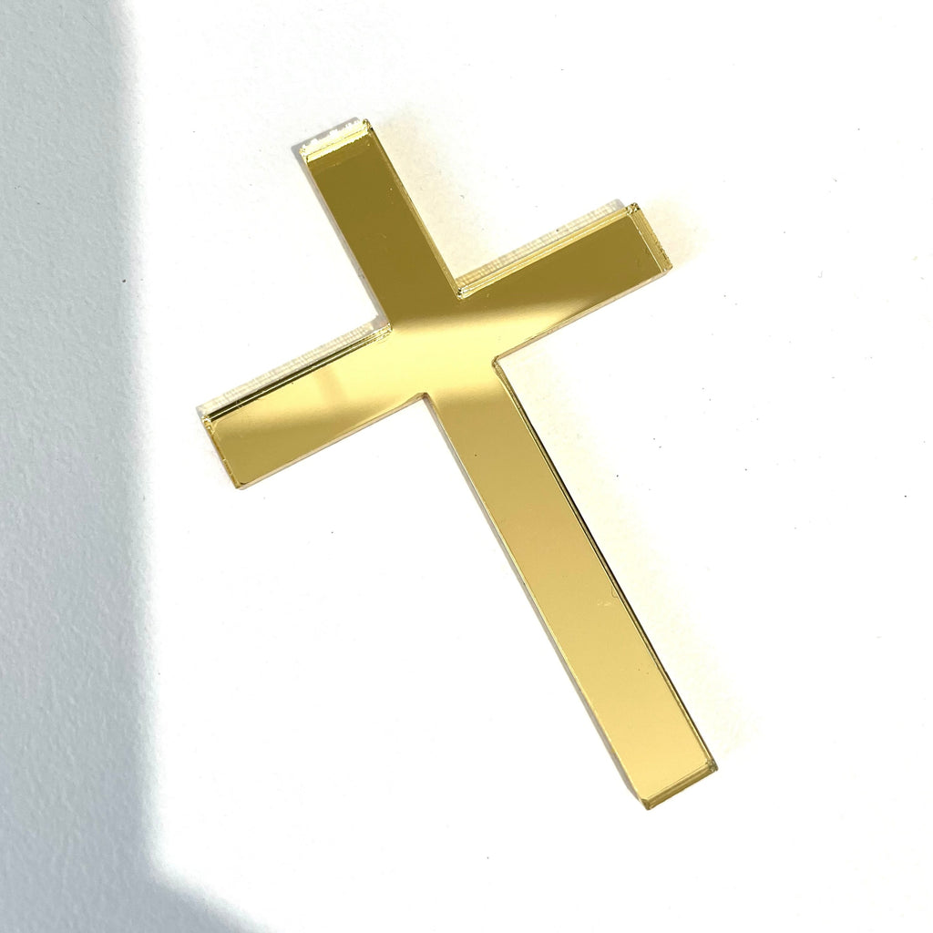 A Solid Cross Charm by Etched Design, perfect for a baptism cake decoration, lies flat on a white surface, casting a slight shadow to the left. The sleek and shiny texture of the gold-colored charm reflects light beautifully.