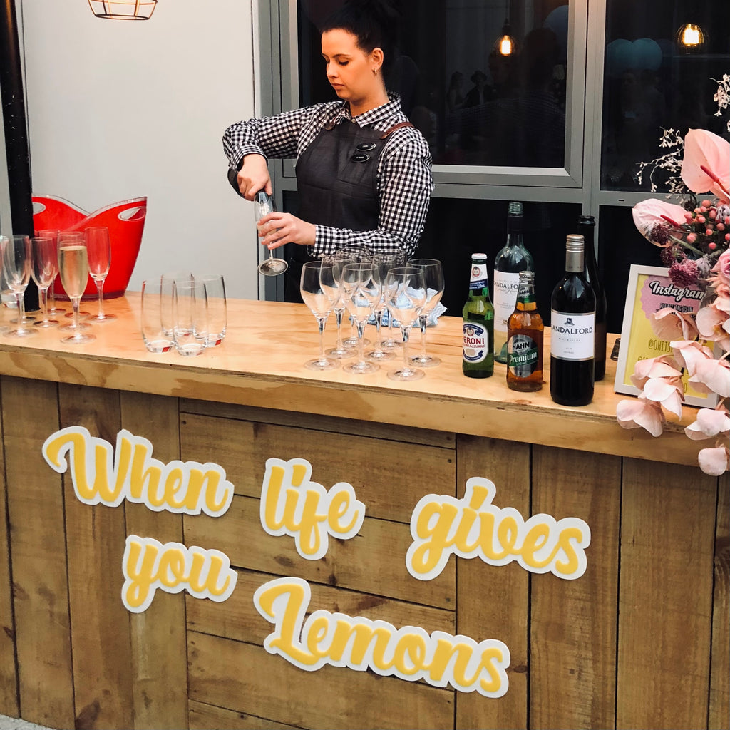 A bartender stands behind a wooden counter with an Etched Design 3D Bar Sign that reads "When life gives you Lemons." She is preparing a drink, surrounded by various alcohol bottles, glasses, and flowers. A die-cut red bucket and a window in the background complete the scene.