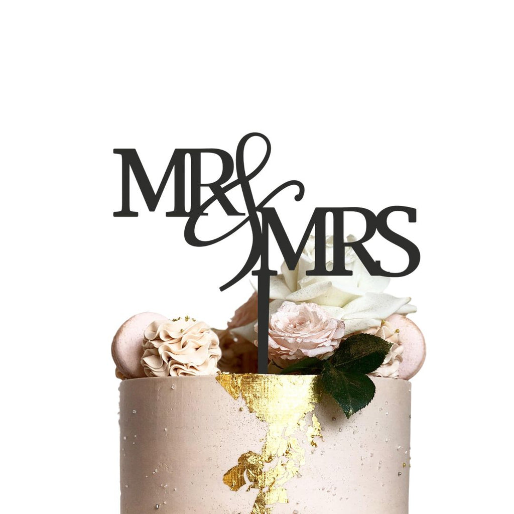 A wedding cake adorned with soft pink and white flowers is topped with the Etched Design "Mr & Mrs" cake topper, which features elegant script. Gold foil accents delicately embellish the white frosting, making the overall design truly enchanting.