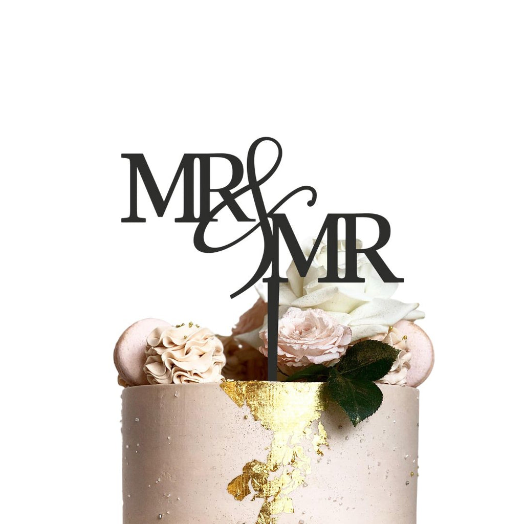 A beige wedding cake adorned with light flowers and macaroons features an Etched Design "Mr & Mr" wooden cake topper in black lettering. The cake boasts gold foil accents and a green leaf for added decoration against a pristine white background.