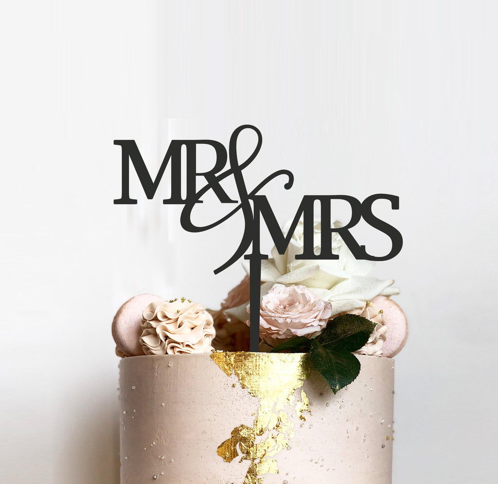 A light pink wedding cake adorned with white and pink roses featuring a bespoke "Mr & Mrs Cake Topper" from Etched Design. Gold leaf embellishments add a touch of elegance to the cake's surface, completing the exquisite wedding cake design.