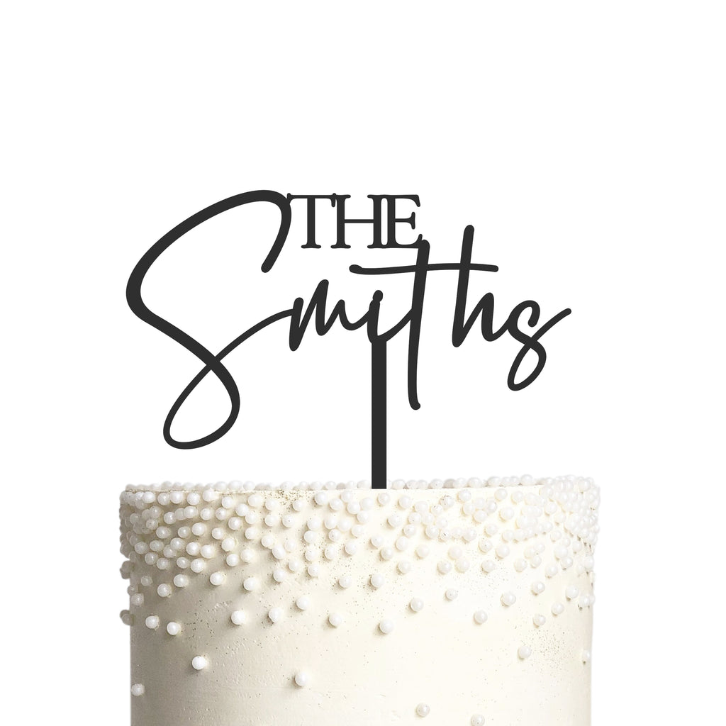 A white cake is adorned with numerous small white pearls, featuring a personalized cake topper from Etched Design's Wedding Surname Cake Topper collection. The topper spells out "The Smiths" in a modern script cursive font against a plain white background.