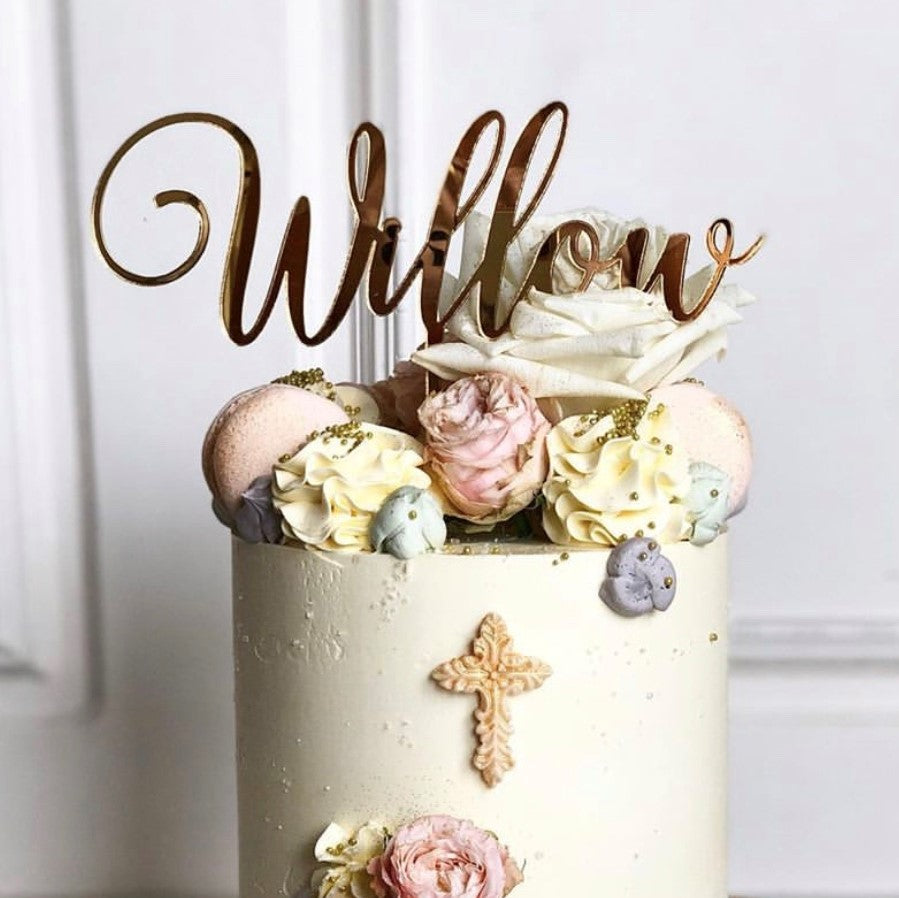 A white, cylindrical cake adorned with various pastel-colored flowers, macarons, and a small cross on its side. A bespoke Name Cake Topper by Etched Design, spelling "Willow," is inserted at the top. The personalized cake decoration is beautifully presented against a white background.