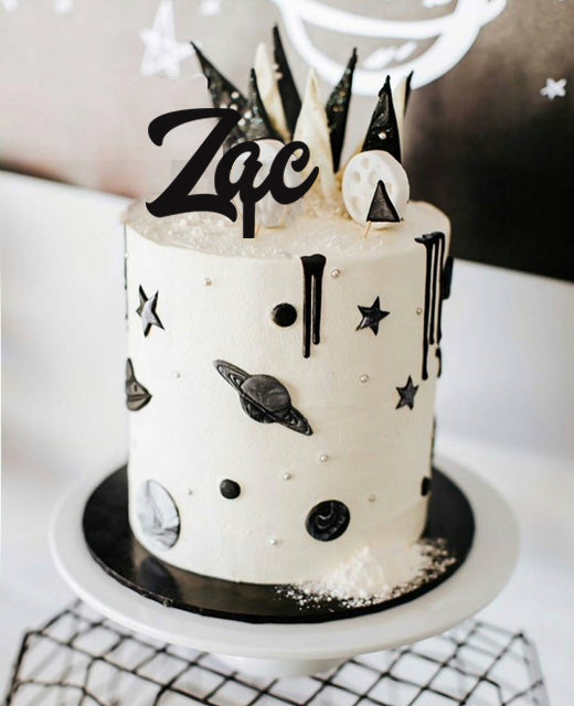 A black and white space-themed cake sits on a stand, decorated with black planets, stars, and rocket ship shapes. The top features an Etched Design Name Cake Topper with "Zac" written in cursive. The background showcases a partial mural of a smiling moon.