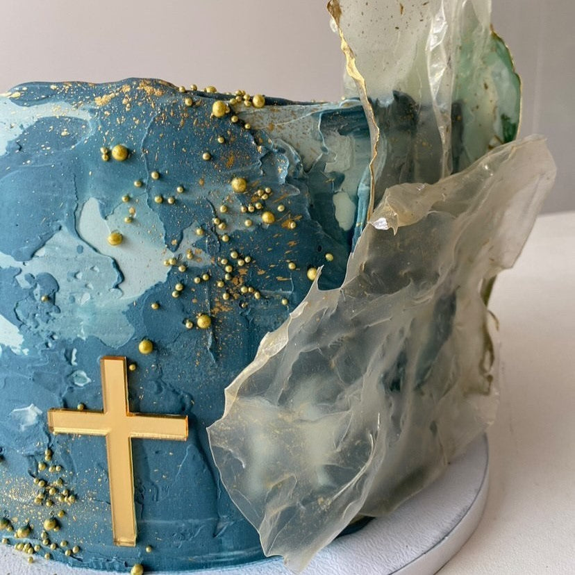 A blue textured cake adorned with Etched Design's Solid Cross Charm, gold beads, and abstract clear sugar sheets, perfect for a baptism or confirmation celebration. This cake boasts a modern and artistic appearance with its combination of blue hues, gold accents, and translucent decorations.