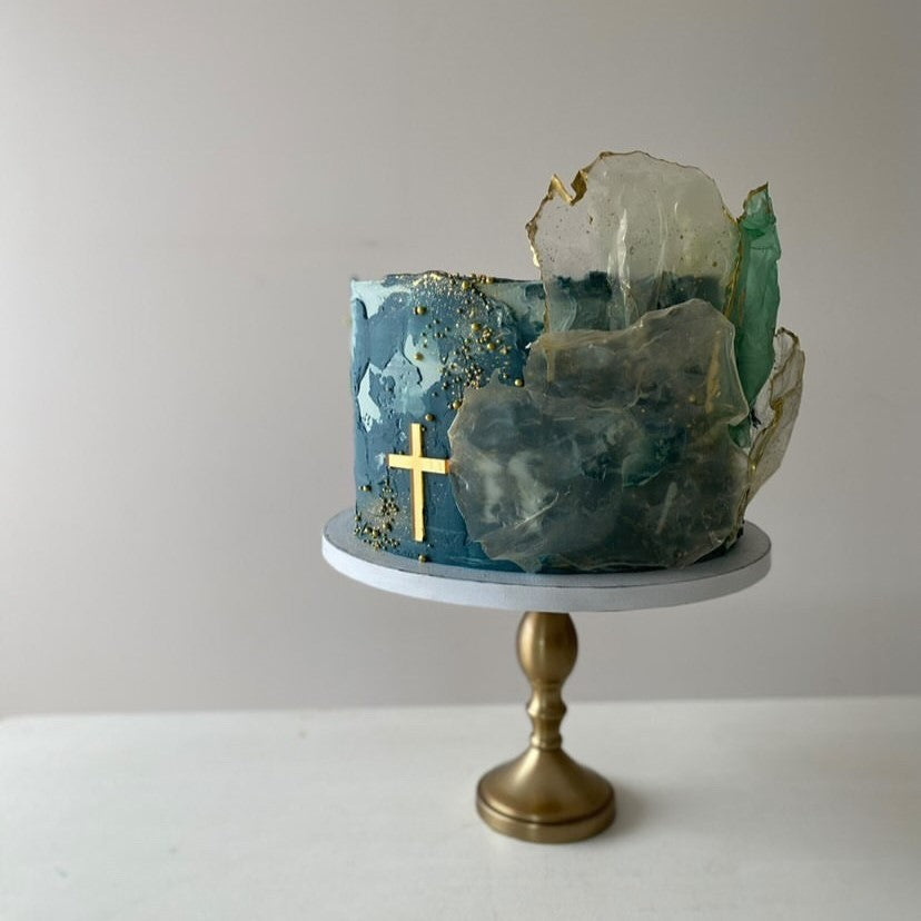 A blue-toned cake adorned with Etched Design's Solid Cross Charm and artistic clear sugar sculptures is displayed on a gold stand. Perfect for a baptism, this cake features textured swirls with gold accents and intricate, translucent sugar decorations resembling crystals. The background is plain and neutral.