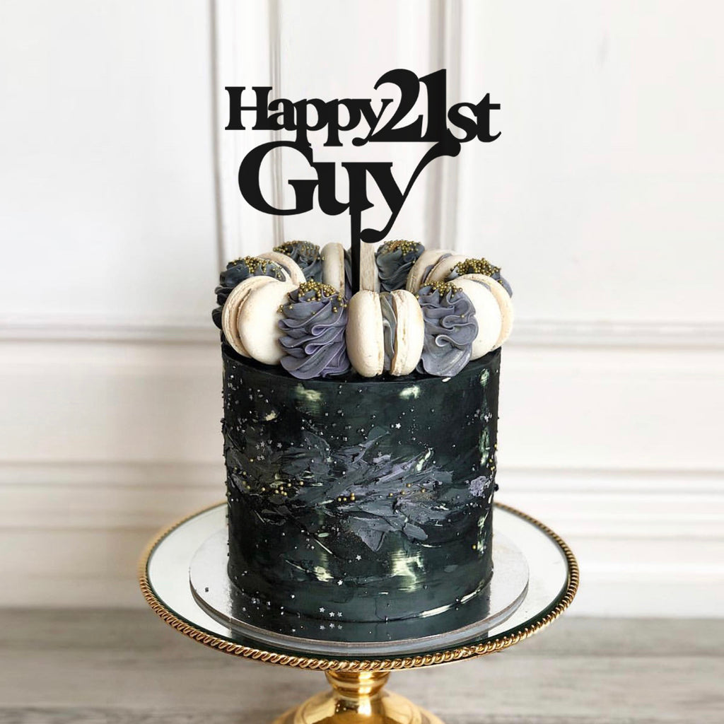 A decorative cake with a dark, elegant design is topped with macarons and a personalized cake topper from Etched Design that reads "Happy 21st Guy." The "Happy Custom Name & Age | Guy" cake sits on a gold-rimmed, reflective cake stand against a white paneled background.