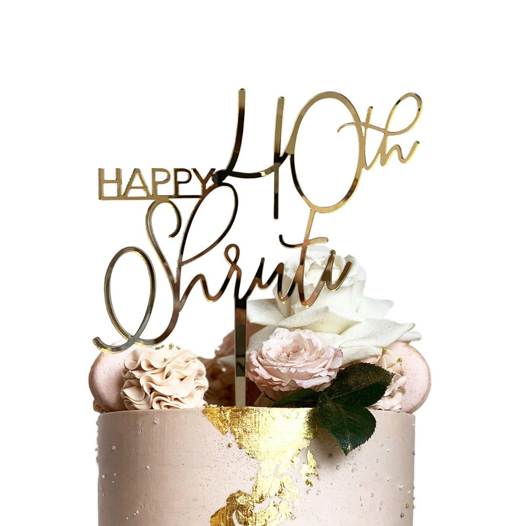 A decorative birthday cake adorned with white roses, pink flowers, and a gold "Happy 40th Shruti" personalized Script Birthday Cake Topper by Etched Design. The cake features gold leaf accents and has a pinkish hue.